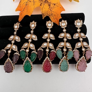 May include: A collection of gold-toned, leaf-shaped earrings with various colored gemstone drops. Each earring features a floral design at the top, with clear crystals and a teardrop-shaped gemstone in shades of purple, green, and red. The earrings are displayed on a black surface.