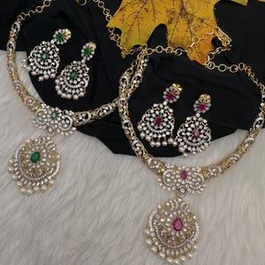 May include: Two gold-toned necklace and earring sets. Each set features a necklace with a large pendant and matching earrings. One set has green gemstones, the other has red gemstones, both with white pearls and clear crystals.