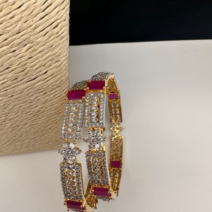 May include: Two gold-toned bangles with clear and red gemstones. The bangles have a rectangular design with clear stones and red rectangular stones. The bangles are on a white surface.