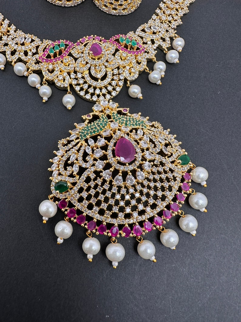 Op de afbeelding: Close-up van een goudkleurige sieradenset met een ketting en hanger. De ketting en hanger zijn versierd met heldere kristallen, robijnkleurige stenen en groene accenten. Witte, parelachtige kralen hangen aan de ketting en hanger.