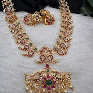 May include: Gold-toned necklace and earrings set with intricate detailing. The necklace features a pendant with red and green gemstones, surrounded by clear crystals and dangling pearls. The earrings have a similar design. The jewelry is displayed on a white, textured surface.