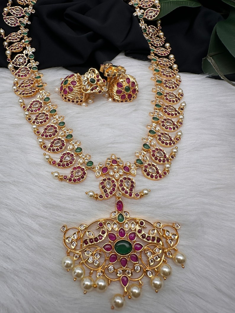 May include: Gold-toned necklace and earrings set with intricate detailing. The necklace features a pendant with red and green gemstones, surrounded by clear crystals and dangling pearls. The earrings have a similar design. The jewelry is displayed on a white, textured surface.