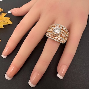 May include: A large, gold-toned ring with a central, round, clear gemstone. The ring is encrusted with smaller, clear gemstones and has a detailed, ornate design. The ring is displayed on a hand with French-manicured nails.