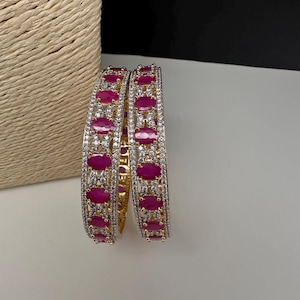 May include: Two gold-tone bangles featuring a central row of oval-shaped, vibrant pink stones. The bangles are embellished with numerous small, clear, sparkling stones set in a metallic frame that surrounds the larger pink oval accents.