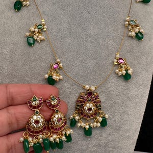 May include: A gold-toned necklace and matching earrings set. The necklace features a central pendant with red and green accents, surrounded by pearls and green beads. The earrings mirror the pendant design, with dangling green beads.