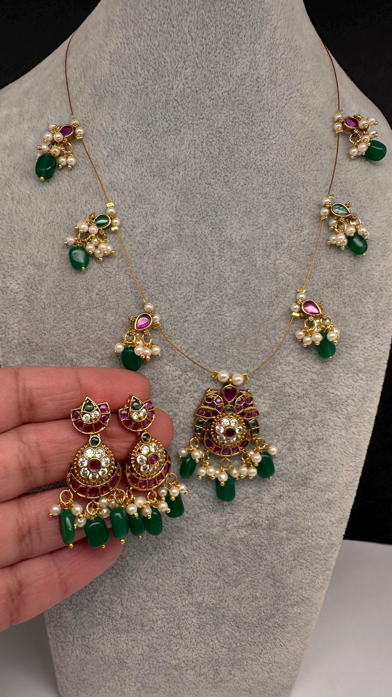 May include: A gold-toned necklace and matching earrings set. The necklace features a central pendant with red and green accents, surrounded by pearls and green beads. The earrings mirror the pendant design, with dangling green beads.