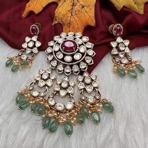 May include: A jewelry set featuring a pendant and earrings. The pendant has a large, oval, red gemstone surrounded by smaller clear stones. The earrings and pendant have a gold-colored metal base with clear stones and green teardrop-shaped beads.