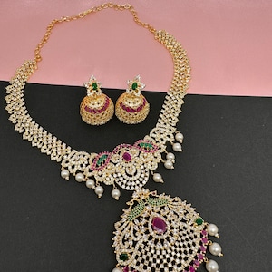 Op de afbeelding: Een goudkleurige ketting- en oorbellenset. De ketting heeft een hanger met een grote, ovale, rode edelsteen, omringd door kleinere witte stenen en groene en rode accenten. De oorbellen zijn rond met vergelijkbare details. Witte parels hangen aan de ketting.