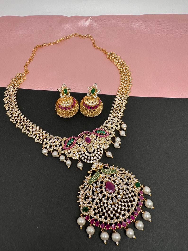 Op de afbeelding: Een goudkleurige ketting- en oorbellenset. De ketting heeft een hanger met een grote, ovale, rode edelsteen, omringd door kleinere witte stenen en groene en rode accenten. De oorbellen zijn rond met vergelijkbare details. Witte parels hangen aan de ketting.