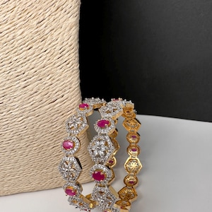 May include: Three gold and silver bangles with red and clear gemstones. The bangles have a geometric design with alternating red oval stones and clear, round stones. The bangles are on a white surface with a woven beige background.