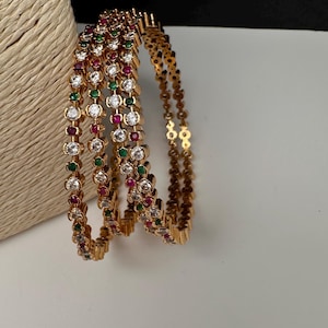 May include: Gold-toned bangles with alternating clear, red, and green gemstones. The bangles are stacked, showcasing their intricate design and sparkle. The jewelry is displayed on a white surface.
