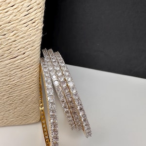 May include: A collection of gold and silver bangles. The gold bangle is on the left, with a row of clear, round stones. The silver bangles are stacked to the right, also with clear, round stones. The bangles are next to a woven basket.