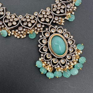 May include: An ornate necklace and earring set featuring a large oval turquoise-colored stone pendant. The jewelry has a silver and gold-tone metal setting with clear, sparkling accents and small, round, turquoise-colored beads. The background is a dark gray.