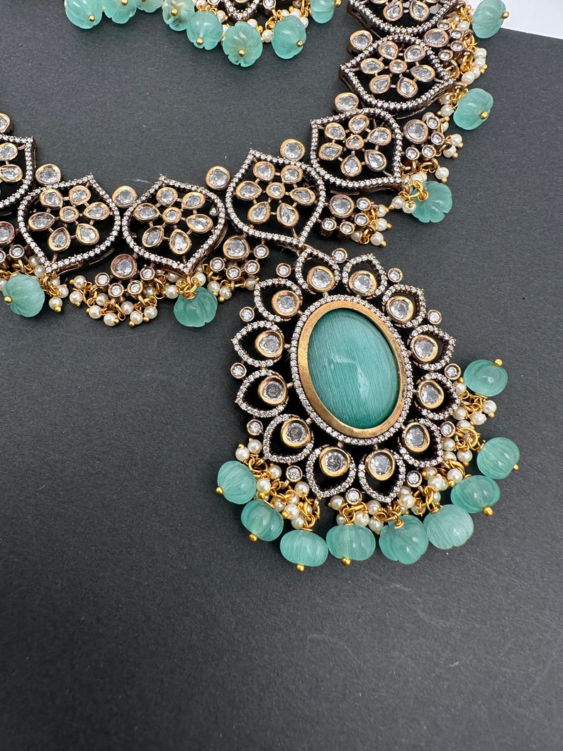 May include: An ornate necklace and earring set featuring a large oval turquoise-colored stone pendant. The jewelry has a silver and gold-tone metal setting with clear, sparkling accents and small, round, turquoise-colored beads. The background is a dark gray.