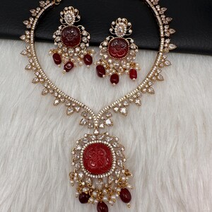 May include: A gold-coloured necklace and earring set. The necklace features a large, round, red pendant with a floral design, surrounded by clear crystals. The earrings mirror the pendant design. Both pieces have dangling red beads and small white pearls.