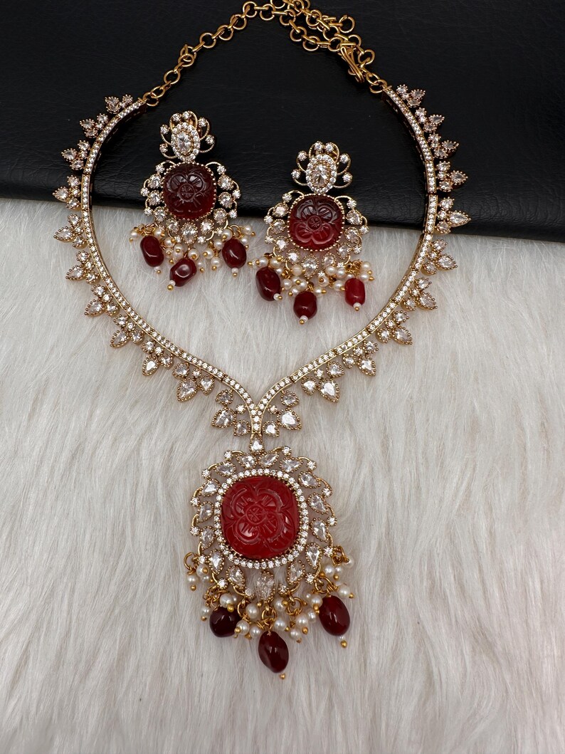 May include: A gold-coloured necklace and earring set. The necklace features a large, round, red pendant with a floral design, surrounded by clear crystals. The earrings mirror the pendant design. Both pieces have dangling red beads and small white pearls.
