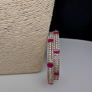 May include: Two gold bangles set with clear and ruby-coloured rectangular stones. The bangles are displayed on a white surface with a woven beige background and a black backdrop.