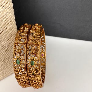 May include: Two gold-colored bangles with intricate floral and figure designs. The bangles feature clear and green gemstones, with a detailed filigree pattern. The background is white and black, highlighting the ornate details of the jewelry.