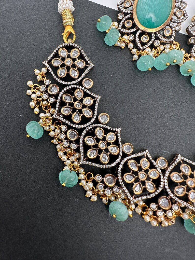 May include: An ornate necklace and earring set. The necklace features a floral design with clear stones set in a black and silver frame, accented with small pearls and light green beads. The earrings have a large oval green stone.