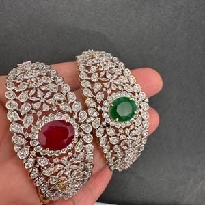 May include: Two elaborate bracelets, one with a large oval ruby-red stone and the other with a green emerald. Both feature a gold base and are encrusted with clear, sparkling crystals in a floral design.