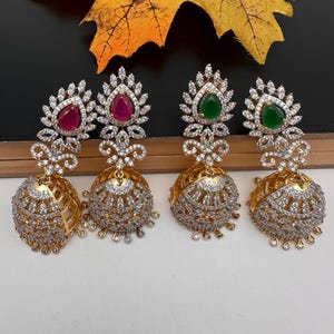 May include: Four gold-toned, ornate earrings with a jhumka design. Each earring features a teardrop-shaped gemstone in red or green, surrounded by clear crystals. The lower portion is a bell shape, also encrusted with crystals.