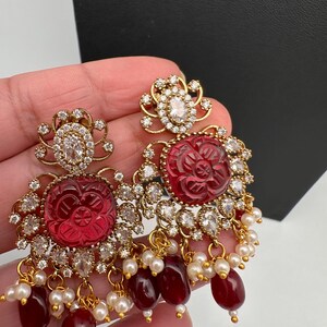 May include: Ornate gold-coloured earrings with red carved floral centres, encircled by clear crystals. The earrings have dangling red beads and small white pearls. The design is elegant.