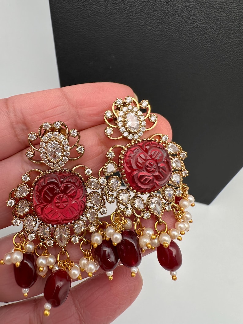 May include: Ornate gold-coloured earrings with red carved floral centres, encircled by clear crystals. The earrings have dangling red beads and small white pearls. The design is elegant.