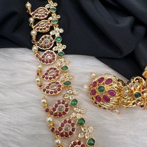 May include: Gold-toned necklace and earring set featuring intricate paisley-shaped designs. The jewelry is adorned with red, green, and clear gemstones, along with small pearls. The set is displayed on a white surface with a black backdrop.