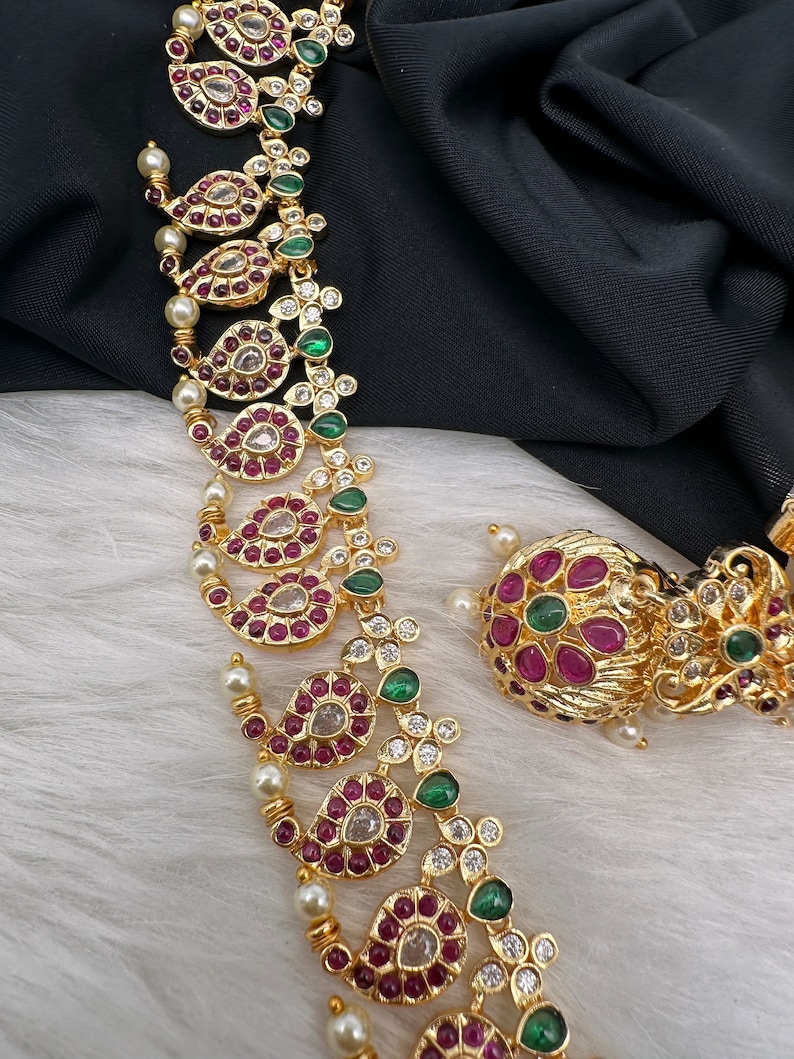 May include: Gold-toned necklace and earring set featuring intricate paisley-shaped designs. The jewelry is adorned with red, green, and clear gemstones, along with small pearls. The set is displayed on a white surface with a black backdrop.
