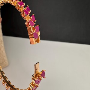 Puede incluir: Una pulsera r&iacute;gida articulada de tono dorado que presenta una serie de piedras fucsia vibrantes en forma de l&aacute;grima dispuestas en una fila continua. La pulsera se muestra en posici&oacute;n abierta, revelando el mecanismo de cierre met&aacute;lico.