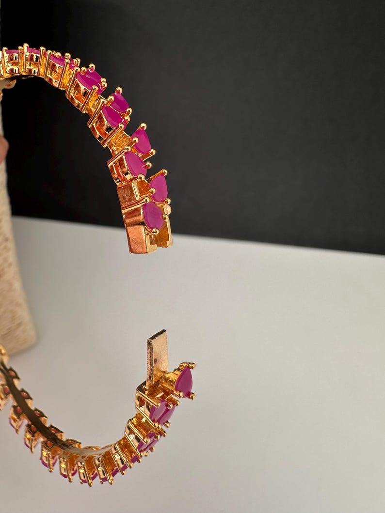 Puede incluir: Una pulsera r&iacute;gida articulada de tono dorado que presenta una serie de piedras fucsia vibrantes en forma de l&aacute;grima dispuestas en una fila continua. La pulsera se muestra en posici&oacute;n abierta, revelando el mecanismo de cierre met&aacute;lico.