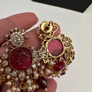 May include: Gold earrings with ruby-red centre stones and clear crystals. The earrings feature a floral design with dangling ruby-red and pearl accents. The earrings are held in a hand.