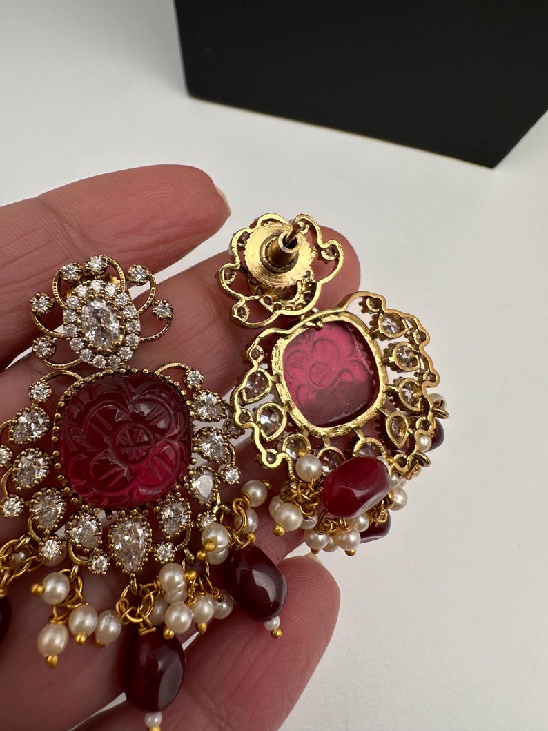 May include: Gold earrings with ruby-red centre stones and clear crystals. The earrings feature a floral design with dangling ruby-red and pearl accents. The earrings are held in a hand.