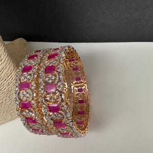 May include: Gold-coloured bangles with alternating ruby-coloured and clear crystal accents. The bangles are displayed on a white surface with a neutral background. The jewellery features a repeating rectangular design.