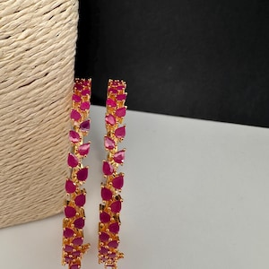 Puede incluir: Dos brazaletes dorados con una hilera continua de piedras de color rosa vibrante en forma de l&aacute;grima. El engaste es de metal dorado pulido y cada piedra est&aacute; sujeta de forma segura con garras, creando un patr&oacute;n uniforme alrededor del aro.