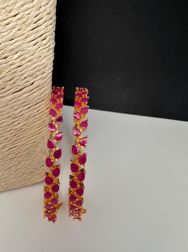 Puede incluir: Dos brazaletes dorados con una hilera continua de piedras de color rosa vibrante en forma de l&aacute;grima. El engaste es de metal dorado pulido y cada piedra est&aacute; sujeta de forma segura con garras, creando un patr&oacute;n uniforme alrededor del aro.