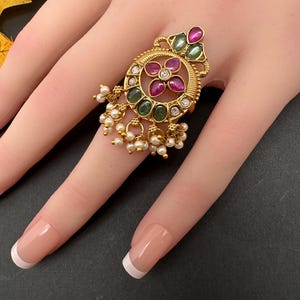 May include: Gold-toned ring with a floral design, featuring red and green gemstones and small white pearls. The ring is ornate and detailed, with a circular centerpiece and dangling pearl accents. The ring is on a finger with a French manicure.