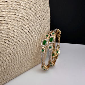 May include: Two gold-coloured bangles with emerald green and clear gemstone accents. The bangles feature square and teardrop-shaped green stones, surrounded by smaller clear stones. The jewellery is displayed on a white surface with a woven beige background.