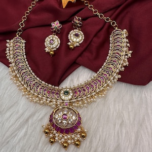 May include: A gold-toned necklace and earring set. The necklace features a series of burgundy-colored, circular accents, interspersed with small pearls. The pendant is a large, circular design with a floral pattern and dangling gold beads. The earrings mirror the design.
