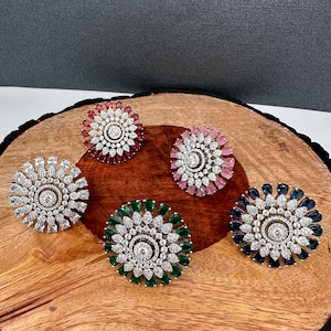 May include: Five flower-shaped rings with a central stone and surrounding petals in various colors: clear, red, pink, green, and blue. The rings are displayed on a wooden surface with a dark gray background.