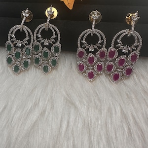 May include: Two pairs of dangling earrings. One pair features emerald green oval-cut stones, and the other pair has ruby red oval-cut stones. Both pairs have a silver-colored, circular design with clear, round accent stones.