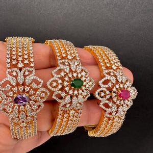 May include: Three gold-toned bangles with intricate floral designs. Each bangle features a central gemstone: purple, green, and red. The bracelets are encrusted with numerous small, clear crystals, creating a sparkling effect.