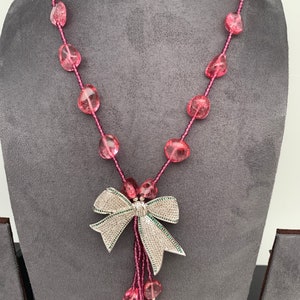 May include: A pink beaded necklace with a silver bow pendant. The necklace features large, irregular pink beads strung on a pink cord. The bow pendant is encrusted with small, clear stones and has a cluster of pink beads hanging below.