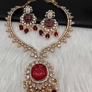 May include: A gold necklace and earring set. The necklace has a large, carved red stone pendant with a floral design, surrounded by clear crystals. The earrings have matching red stone centres and dangling red beads. The set is embellished with small pearls.