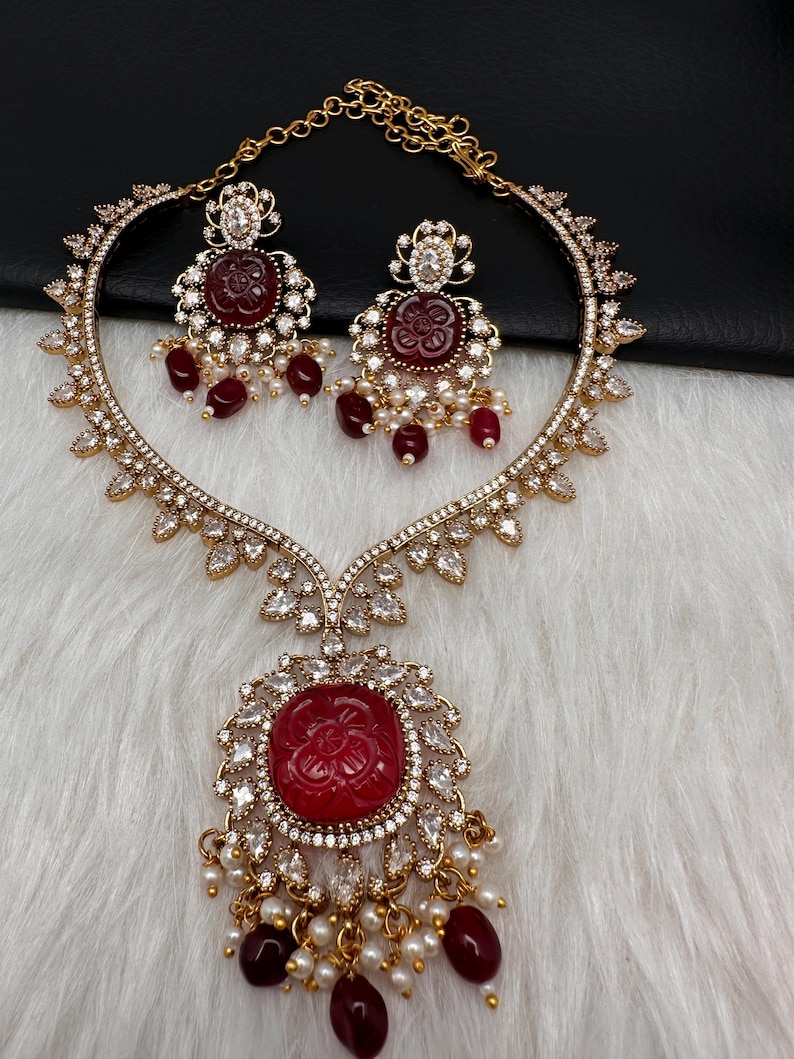 May include: A gold necklace and earring set. The necklace has a large, carved red stone pendant with a floral design, surrounded by clear crystals. The earrings have matching red stone centres and dangling red beads. The set is embellished with small pearls.