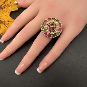 May include: A gold-toned ring with a circular design, featuring alternating red and green gemstones, and small white stones. The ring is set on a hand with a neutral skin tone, with French-manicured nails.