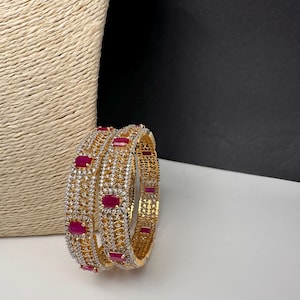 May include: Gold-toned bangles with a repeating pattern of red rectangular stones and clear crystals. The bangles are displayed on a white surface with a textured beige background.
