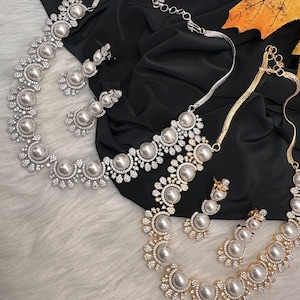 May include: Two matching jewelry sets, each including a necklace and earrings. One set is silver-toned, the other gold-toned. Both feature large, faux pearl accents surrounded by clear, sparkling stones. The jewelry rests on a black fabric and white fur.
