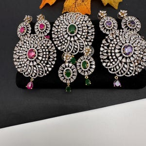 May include: A collection of ornate, sparkling jewelry sets. Each set includes a large pendant and matching earrings. The earrings and pendants feature a central gemstone, surrounded by smaller, clear stones. The gemstones are pink, green, and purple.