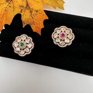 May include: Two floral-shaped stud earrings. Each earring features a central gemstone, one green and one pink, surrounded by clear stones and a gold-colored metal frame. The earrings are displayed on a black surface with a fall leaf in the background.